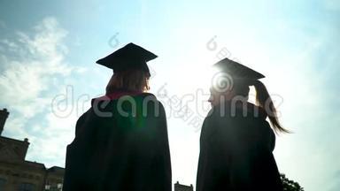 后景两个快乐的学生在壁炉前庆祝他们的毕业在大学大楼前。 新毕业